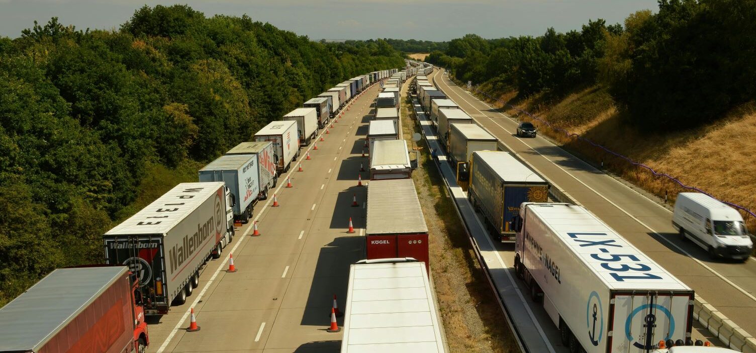 A long queue of heavy goods vehicles (HGVs) lined up on a motorway, separated by traffic cones.